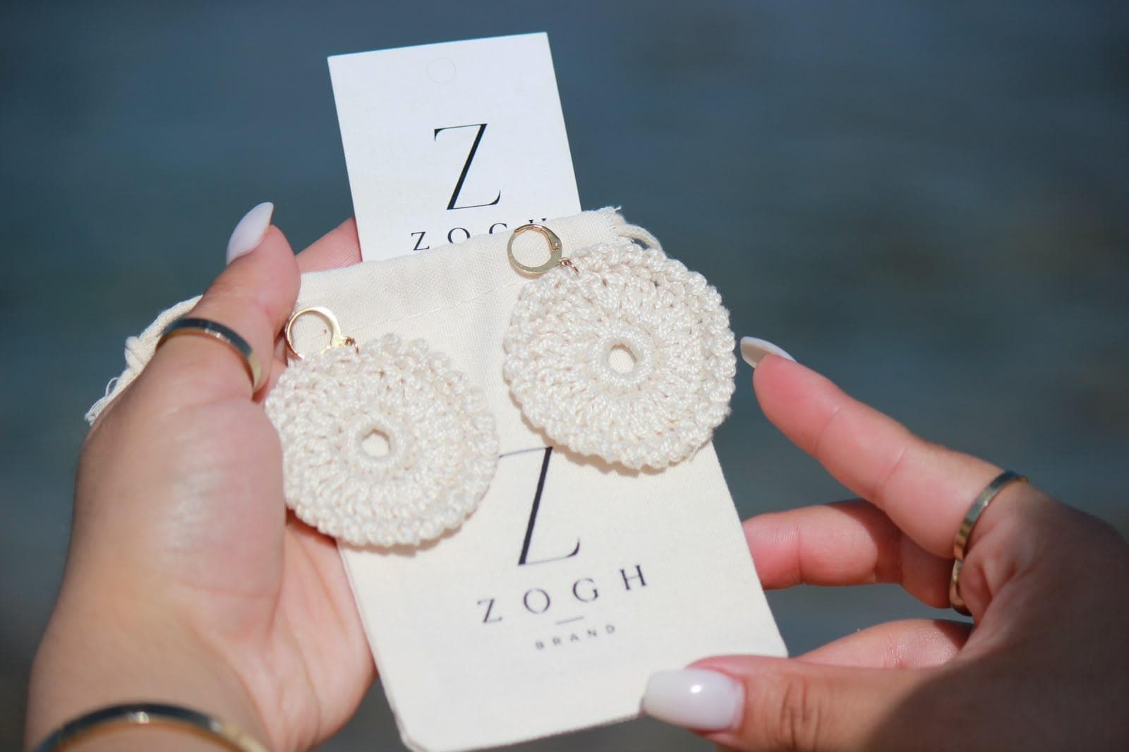 Pendientes de crochet blancos sobre la tarjeta y la bolsita de tela con el logo ZoghBrand, frente al mar